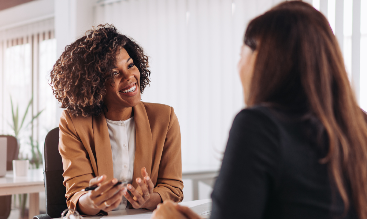 Black woman wearing natural curly shoulder length hair, a tan blazer and white blouse smiling at a white woman wearing long brown hair and wearing a black blazer