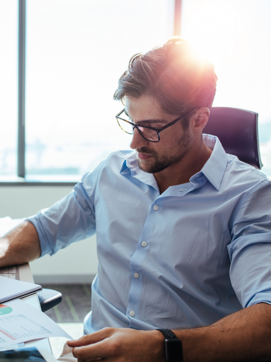 White man with dark wavy short hair and a beard with dark rimmed glasses wearing a light blue collared button-down shirt look down at some documents at a desk with the sun rays hitting the back of his head through a window